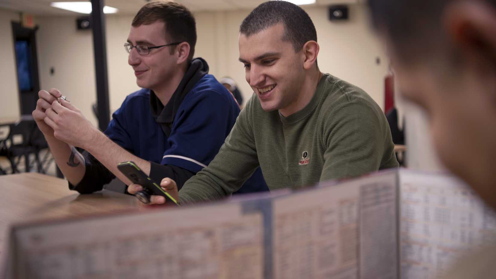 Players smile across a Dungeons & Dragons table; one holds a die and another holds a phone behind a game screen.